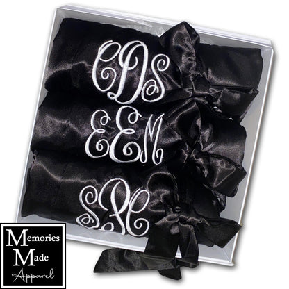 The photo displays three black satin robes with a monogram stitched out on the front left breast. The monograms are white. The robes are displayed in a row. They are rolled up and tied with the robes tie. The background is white. The logo for  Memories Made Apparel is in the lower left corner.