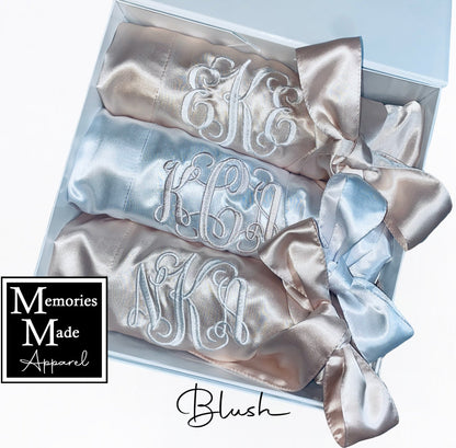 Three robes with custom embroidered monograms on the left chest. Two robes are blush, one robe is white. Each robe is rolled and tied with the robes satin belt. The robes are displayed in a white box against a white background. The logo for Memories Made Apparel is in the lower left corner. The word blush is in the lower right hand corner. 