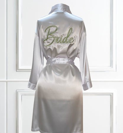 White satin robe on a mannequin. The view is the backside of the robe. Bride is embroidered on the backside in the color sage. The background is a white wall with box trim. 