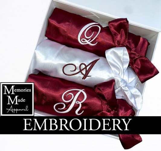 Two burgundy satin robes and one white satin robe with custom embroidered single initials on the left chest. . Each robe is rolled and tied with the matching robe belt. The robes are displayed in a white box against a white background. The logo for Memories Made Apparel is in the lower left side. The word "embroidery" is on a banner in the lower portion of the picture. 