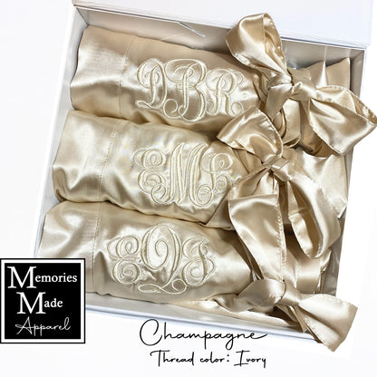 The photo displays three champagne color satin robes with a monogram stitched out on the front left breast. The monograms are in the color ivory. The robes are displayed in a row inside of a white gift box. They are rolled up and tied with the robes tie. The background is white. The words "champagne"  and "Thread color: Ivory"  are in the lower center of the photo. The logo for  Memories Made Apparel is in the lower left corner. 