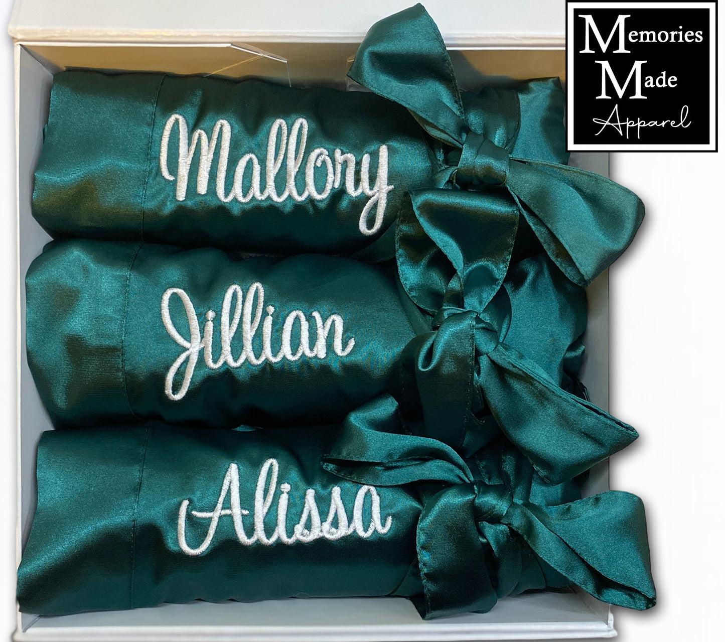 3 Green robes with custom embroidery on the left chest it the thread color white. Each robe has a custom name embroidered on the left chest. Each robe is rolled and tied with the matching satin belt. The three robes are displayed in a white box against a white background. The logo for Memories Made Apparel is in the upper right corner of the pic. 