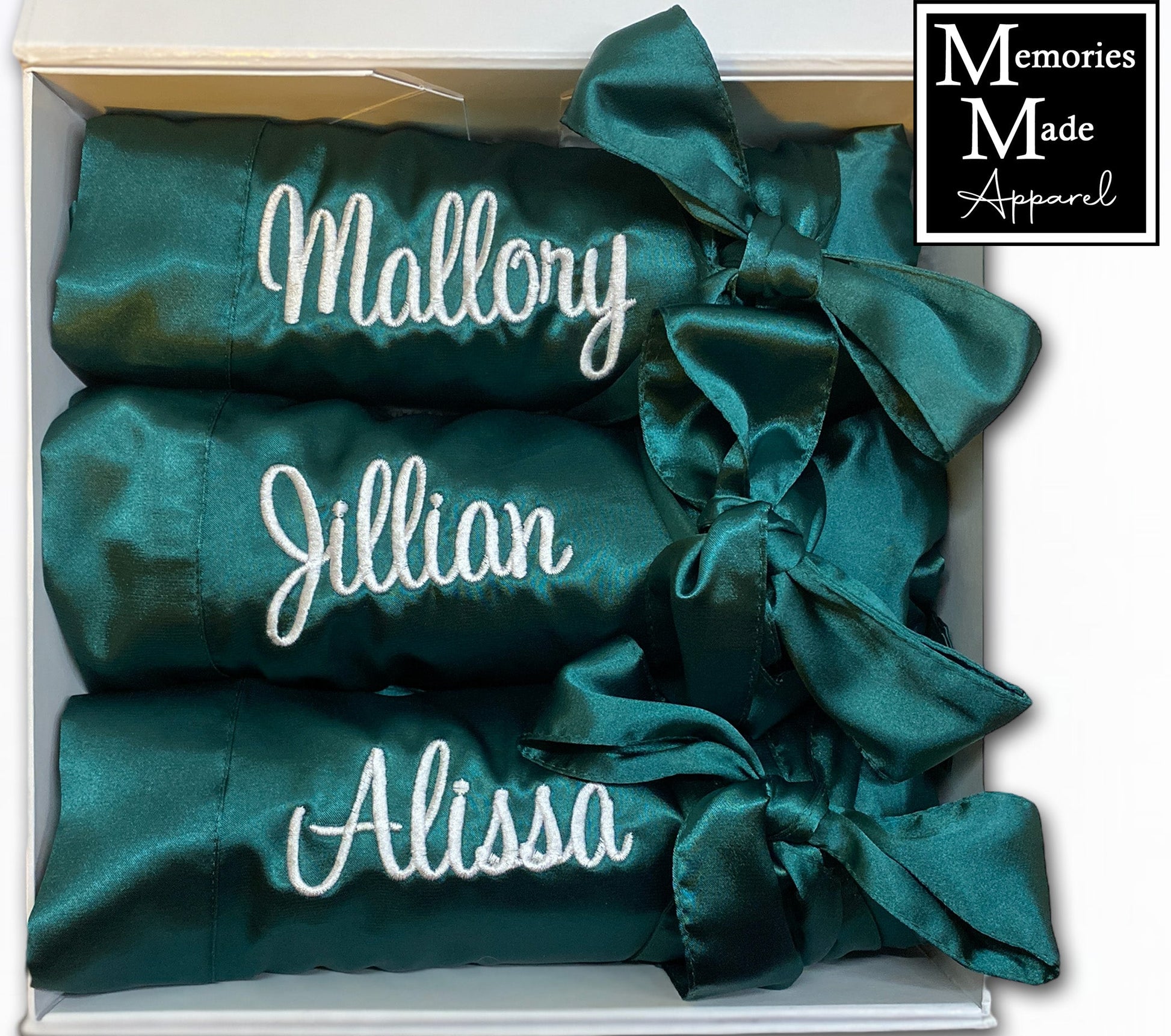 3 Green robes with custom embroidery on the left chest it the thread color white. Each robe has a custom name embroidered on the left chest. Each robe is rolled and tied with the matching satin belt. The three robes are displayed in a white box against a white background. The logo for Memories Made Apparel is in the upper right corner of the pic. 