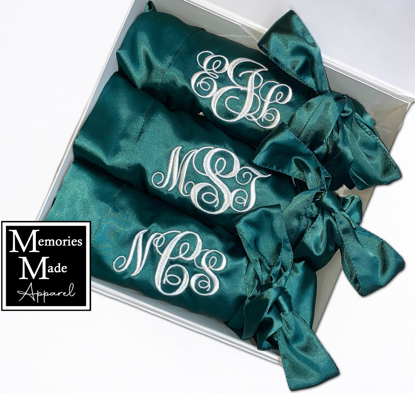 3 Green satin robes with custom embroidery on the left breast in white thread. Each robe has a 3 letter monogram. The robes are rolled and tied with the satin belt. The robes are displayed in a white box and are against a white background. The logo for Memories Made Apparel is in the lower left corner. 