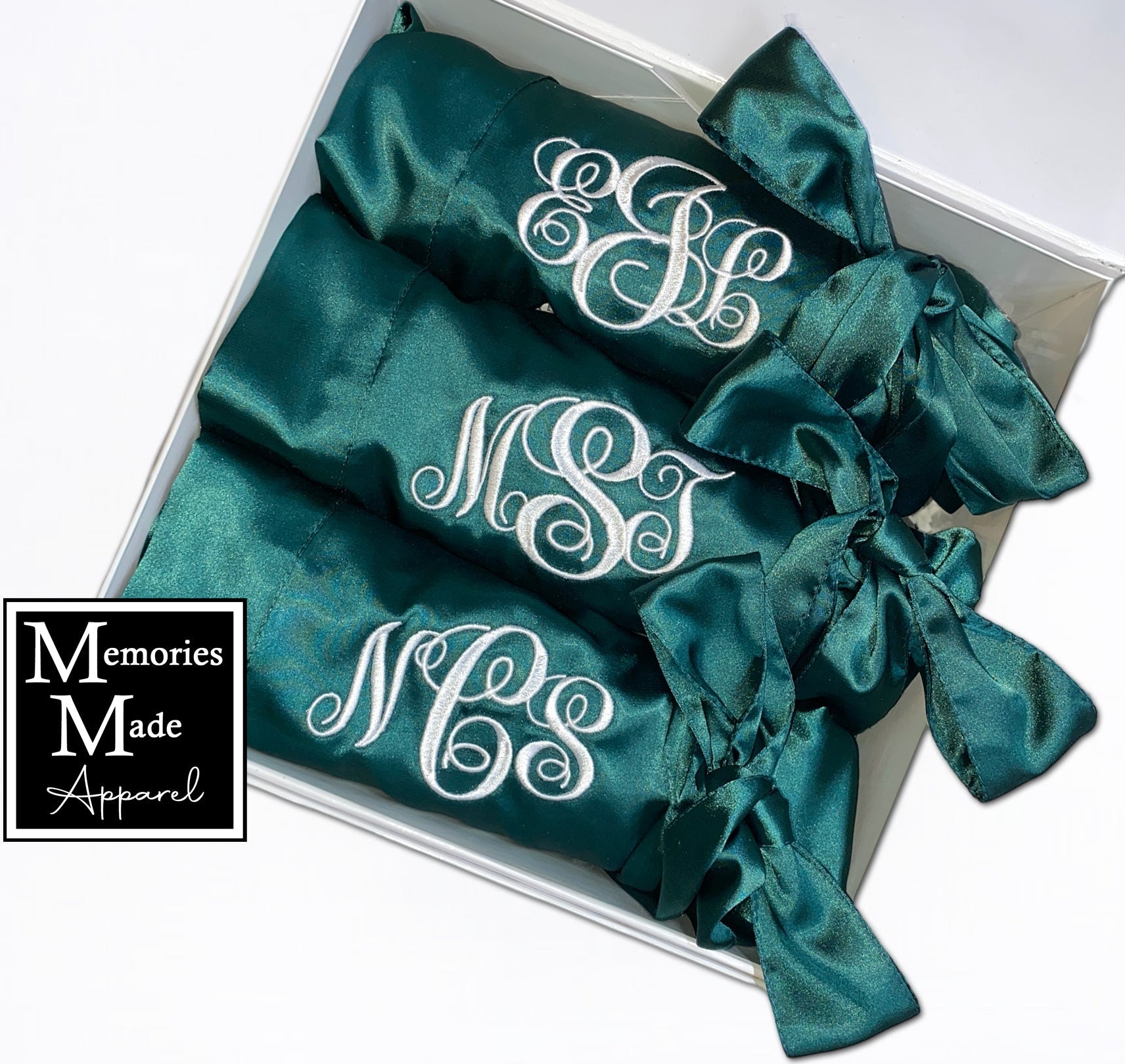 3 Green satin robes with custom embroidery on the left breast in white thread. Each robe has a 3 letter monogram. The robes are rolled and tied with the satin belt. The robes are displayed in a white box and are against a white background. The logo for Memories Made Apparel is in the lower left corner. 