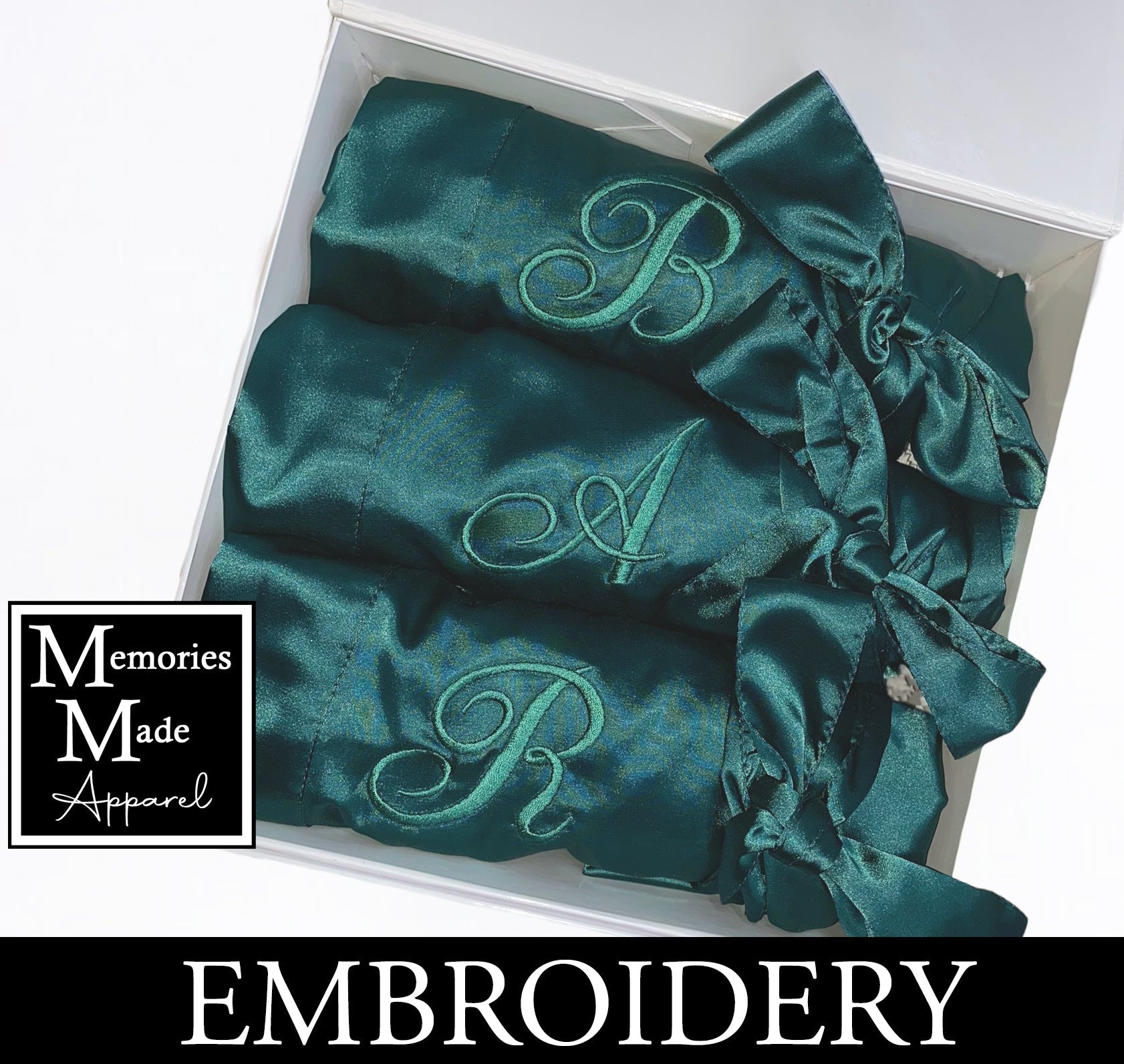 3 Green satin robes with custom embroidery on the left breast. Each robe has a single initial embroidered. Each robe is rolled and tied with the satin robe tie. The robes are displayed in a white box against a white background. The logo for Memories Made Apparel is in the lower left corner. A banner with the word "Embroidery" is across the bottom of the image. 