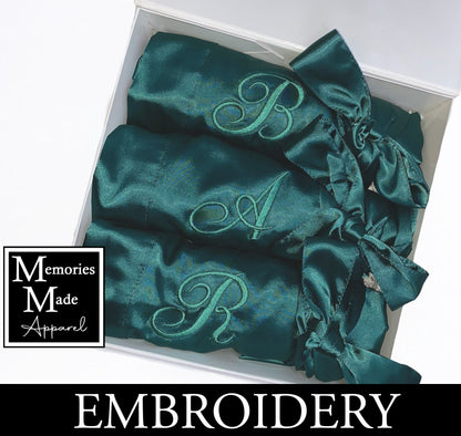 3 Green satin robes with custom embroidery on the left breast. Each robe has a single initial embroidered. Each robe is rolled and tied with the satin robe tie. The robes are displayed in a white box against a white background. The logo for Memories Made Apparel is in the lower left corner. A banner with the word "Embroidery" is across the bottom of the image. 