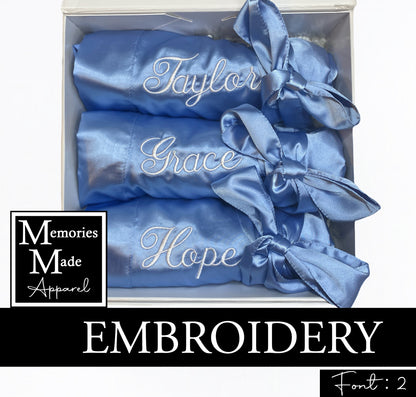 Three light blue satin robes with custom embroidered names on the left chest in the thread color white. The robes are rolled and tied with the matching robe belt. The robes are displayed in a white box against a white background. The logo for Memories Made Apparel is in the lower left side. The word "embroidery" is on a banner in the lower portion of the picture. The words "Font: 2" are in the lower right hand corner. 