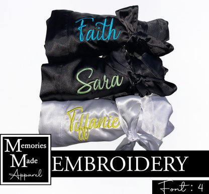 Photo shows three satin robes with names embroidered on the left breast. The robes are rolled up and tied with the robe belt and displayed  in a row. Two of the robes are black. One has a name stitched out in aqua thread. The other has a name stitched out in sage thread. The third robe is white and has a name stitched out in yellow thread. There is a logo for Memories Made Apparel  in the lower left corner. "Font 4" is in the lower right corner. A black banner is across the photo with the word "embroidery".