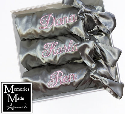 Three gray robes with custom embroidered names on the left chest. Each robe is rolled and tied with the robes satin belt. The robes are displayed in a white box against a white background. The logo for Memories Made Apparel is in the lower left corner.