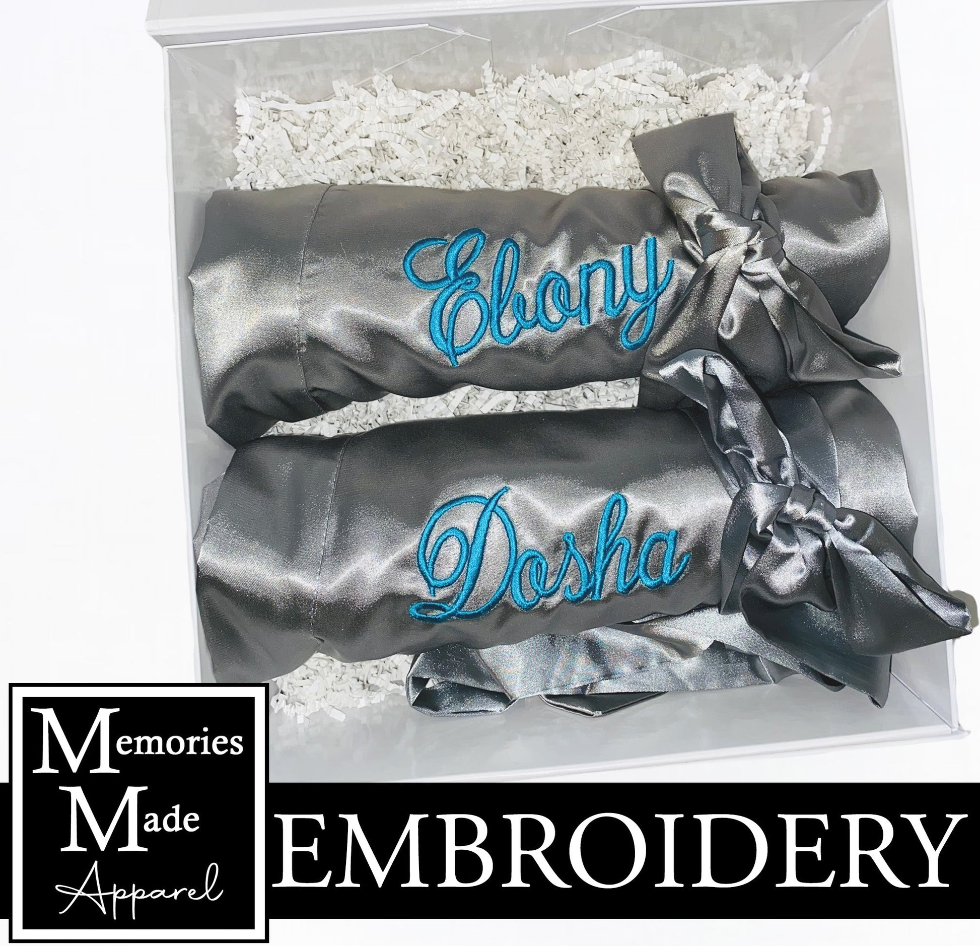 Two gray robes with custom embroidered names on the left chest. Each robe is rolled and tied with the robes satin belt. The robes are displayed in a white box against a white background. The logo for Memories Made Apparel is in the lower left corner. The word "embroidery" is displayed on a banner in the lower portion of the picture. 