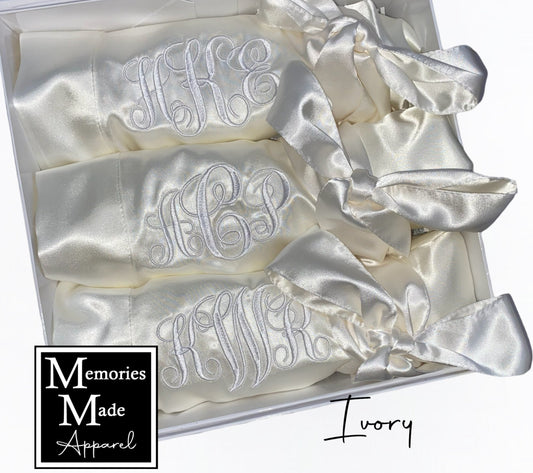 3 satin ivory robes displayed in a white box against a white background. Robes are rolled and tied with the satin robe belt. Each robe features a custom 3 letter monogram in white thread. There is a logo for Memories Made Apparel in the bottom left corner. "Ivory" is in the lower right corner.