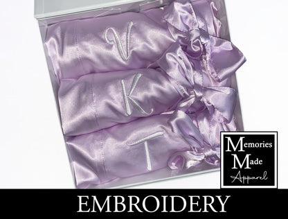 The photo displays three lavender color satin robes with single initials  stitched out on the front left breast. The thread color is white. The robes are displayed in a row. They are rolled up and tied with the robes tie. The background is white. The logo for  Memories Made Apparel is in the lower right corner.