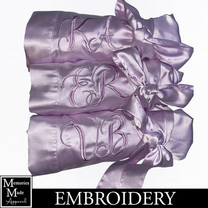The photo displays three lavender color satin robes with two letter monograms  stitched out on the front left breast. The thread color is lavender. The robes are displayed in a row. They are rolled up and tied with the robes tie. The background is white. The logo for  Memories Made Apparel is in the lower left corner.