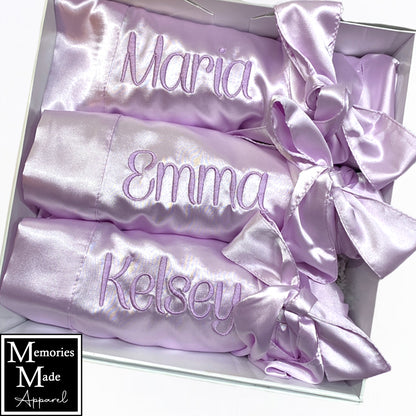 The photo displays three lavender color satin robes with names stitched out on the front left breast. The thread color is  lavender. The robes are displayed in a row. They are rolled up and tied with the robes tie. The background is white. The logo for  Memories Made Apparel is in the lower left corner.
