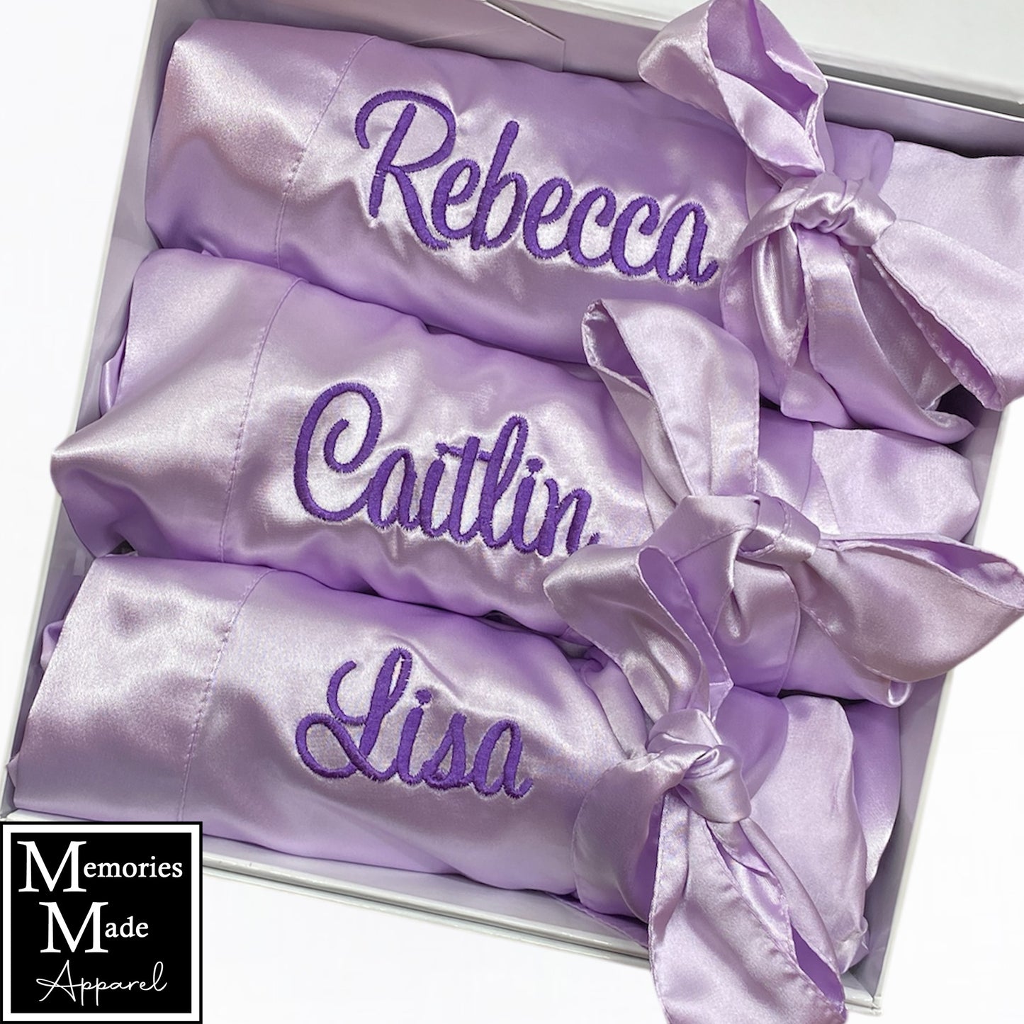 The photo displays three lavender color satin robes with names  stitched out on the front left breast. The thread color is purple. The robes are displayed in a row. They are rolled up and tied with the robes tie. The background is white. The logo for  Memories Made Apparel is in the lower left corner.