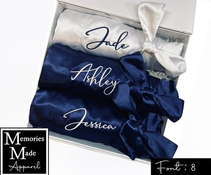 Picture shows three satin robes displayed in a white gift box with custom embroidery on the left breast. The robes are rolled up and tied with the robe belt and displayed  in a row. Two of the robes are in the color navy and have a name embroidered in the color white. One of the robes is white in color and has a name embroidered in font 12 in the color navy. The background is white. Font 8 is in the lower right corner. The logo for  Memories Made Apparel is in the lower left corner. 