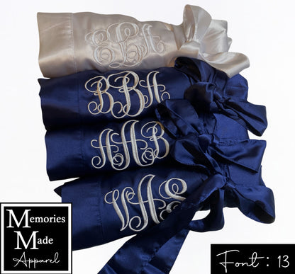 Picture shows four satin robes with custom embroidery on the left breast. The robes are rolled up and tied with the robes belt and displayed  in a row. Three of the robes are in the color navy and have a monogram embroidered in the color ivory. One of the robes the color ivory and has a monogram embroidered in the color navy. The background is white. Font 13 is in the lower right corner. The logo for  Memories Made Apparel is in the lower left corner. 