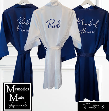 Picture shows three satin robes displayed hanging up against a white background. The view is of the backside. Two of the robes are in the color navy and have a the titles of Brides Maid on one, and of Maid of Honor on the other embroidered in the color navy. One of the robes is white in color and has the title of Bride embroidered on the back in the color navy. The background is white. Font 8 is in the lower right corner. The logo for  Memories Made Apparel is in the lower left corner. 