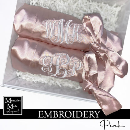 Two pink robes with white custom embroidered monograms on the left chest. Each robe is rolled and tied with the robes satin belt. The robes are displayed against a white background. The logo for Memories Made Apparel is in the lower left corner. A banner with the word "embroidery" is displayed across the bottom. The word pink is in the lower right hand corner.  