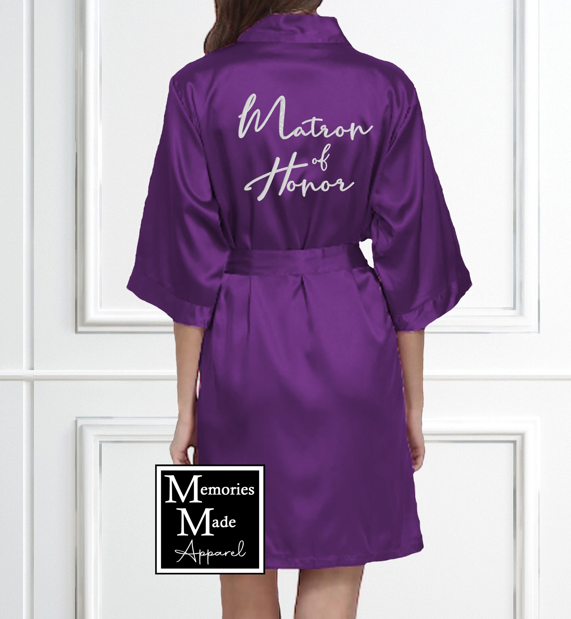 The backside view of a model wearing a purple robe with the words "Maid of Honor" embroidered across the back in white thread. The model is standing against a white wall with white box trim. The logo for Memories Made Apparel is in the lower portion of the picture. 