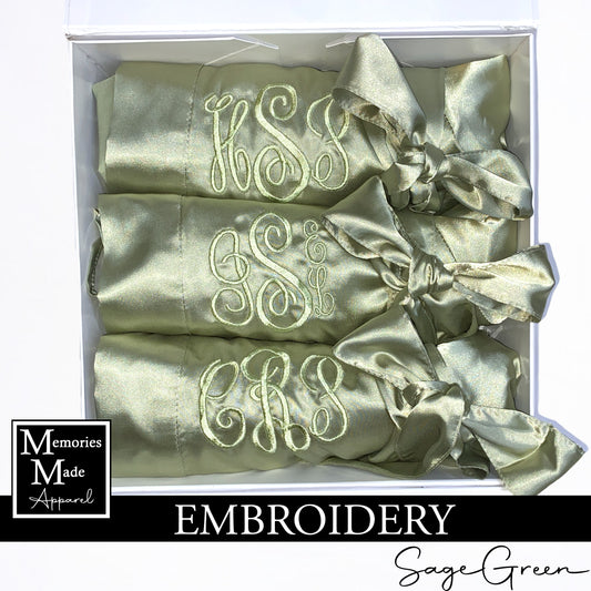 Three sage green satin robes with custom embroidered monograms on the left chest in the thread color sage green. The robes are rolled and tied with the matching robe belt. The robes are displayed in a white box against a white background. The logo for Memories Made Apparel is in the lower left side. The word "embroidery" is on a banner in the lower portion of the picture. The words "Sage Green" are in the lower right hand corner. 