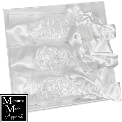 Picture shows three satin robes displayed in a white gift box with custom embroidery on the left breast. The robes are rolled up and tied with the robe belt and displayed  in a row. The three robes are white and have a monogram embroidered in the color white. . The background is white. The logo for  Memories Made Apparel is in the lower left corner. 