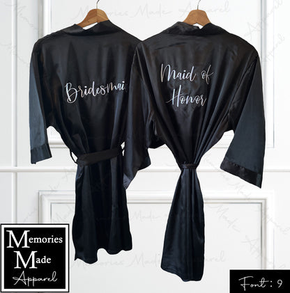 Photo shows two black satin robes hanging up against a white wall with box trim. The view is the back side of the robes. One robe has "Bridesmaid" embroidered across the back in white thread. The other has "Maid of Honor" embroidered across the back in white thread. The logo for  the company Memories Made Apparel is in the lower left corner. "Font: 9" is in the lower right corner. There is a watermark over the photo. 