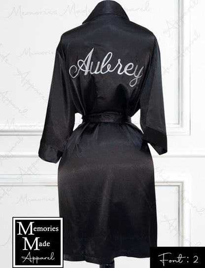 Photo shows a black satin robe displayed on a mannequin. The view is the backside. The name "Aubrey" is embroidered on the back. The background is a white wall with box trim. The logo for Memories Made Apparel is in the lower left corner. "Font: 2" is in the lower right corner. 