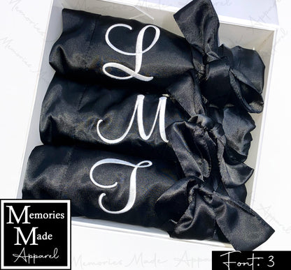 The photo displays three black satin robes with a single initial stitched out on the front left breast. The initials are white. The robes are displayed in a row inside of a white gift box. They are rolled up and tied with the robes tie.  The background is white. The logo for  Memories Made Apparel is in the lower left corner. The word "Font: 3" is in the lower right corner. 