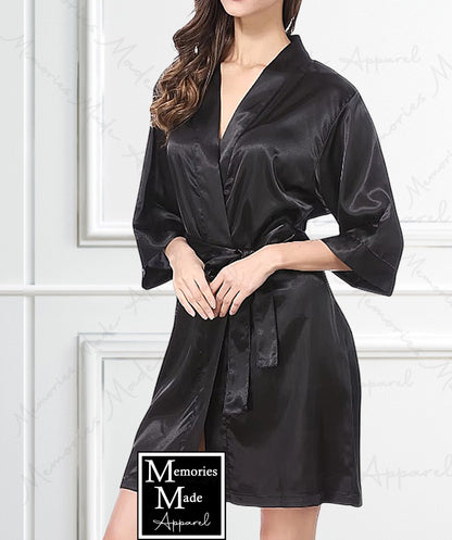 Photo shows a female appearing model wearing a black satin robe. The view is the front side. The model is standing in front of a white wall with box trim. The logo for Memories Made Apparel is on bottom middle of the photo. There is a watermark across the photo. 