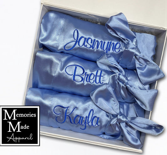 Three light blue satin robes with custom embroidered names on the left chest in the thread color royal blue. The robes are rolled and tied with the matching robe belt. The robes are displayed in a white box against a white background. The logo for Memories Made Apparel is in the lower left side.