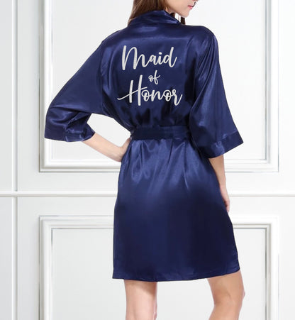 The picture displays the back side view of a woman wearing a navy robe. Maid of Honor is embroidered on the back of the robe in the color white. The background is a white wall with white box trim. 