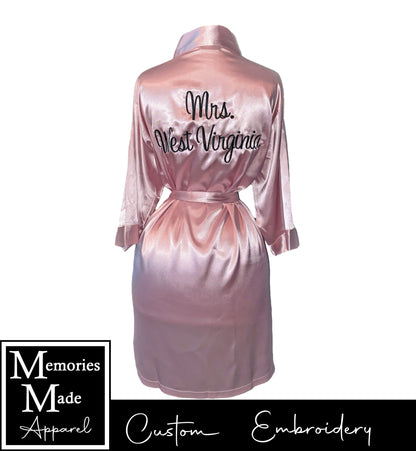 Pink robe on a mannequin with custom embroidery across the back in black thread. on the left chest. The robe is displayed against a white background. The logo for Memories Made Apparel is in the lower left corner. 