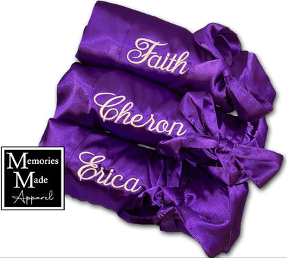 3 purple satin robes with custom embroidery on the left chest in white thread. The robes are rolled and tied with the robes satin tie. The robes are displayed against a white background. The logo for Memories Made Apparel is in the lower left corner. 