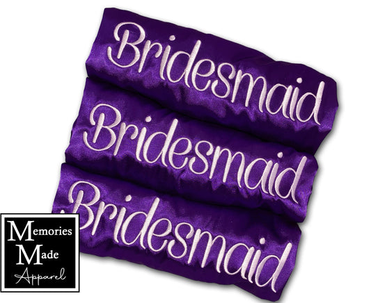 3 Purple robes with embroidery in white  thread. The word "Bridesmaid" is embroidered on each robe. The 3 robes are rolled and displayed against a white background. The logo for Memories Made Apparel is in the lower left hand corner. 